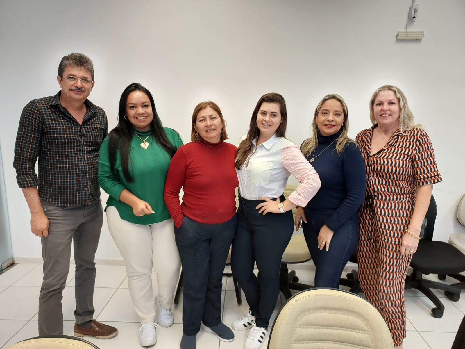 Grupo de seis pessoas posando para foto em ambiente interno, provavelmente de trabalho. Há um homem e cinco mulheres, todas sorrindo. Ao fundo, cadeiras de escritório e parede branca.