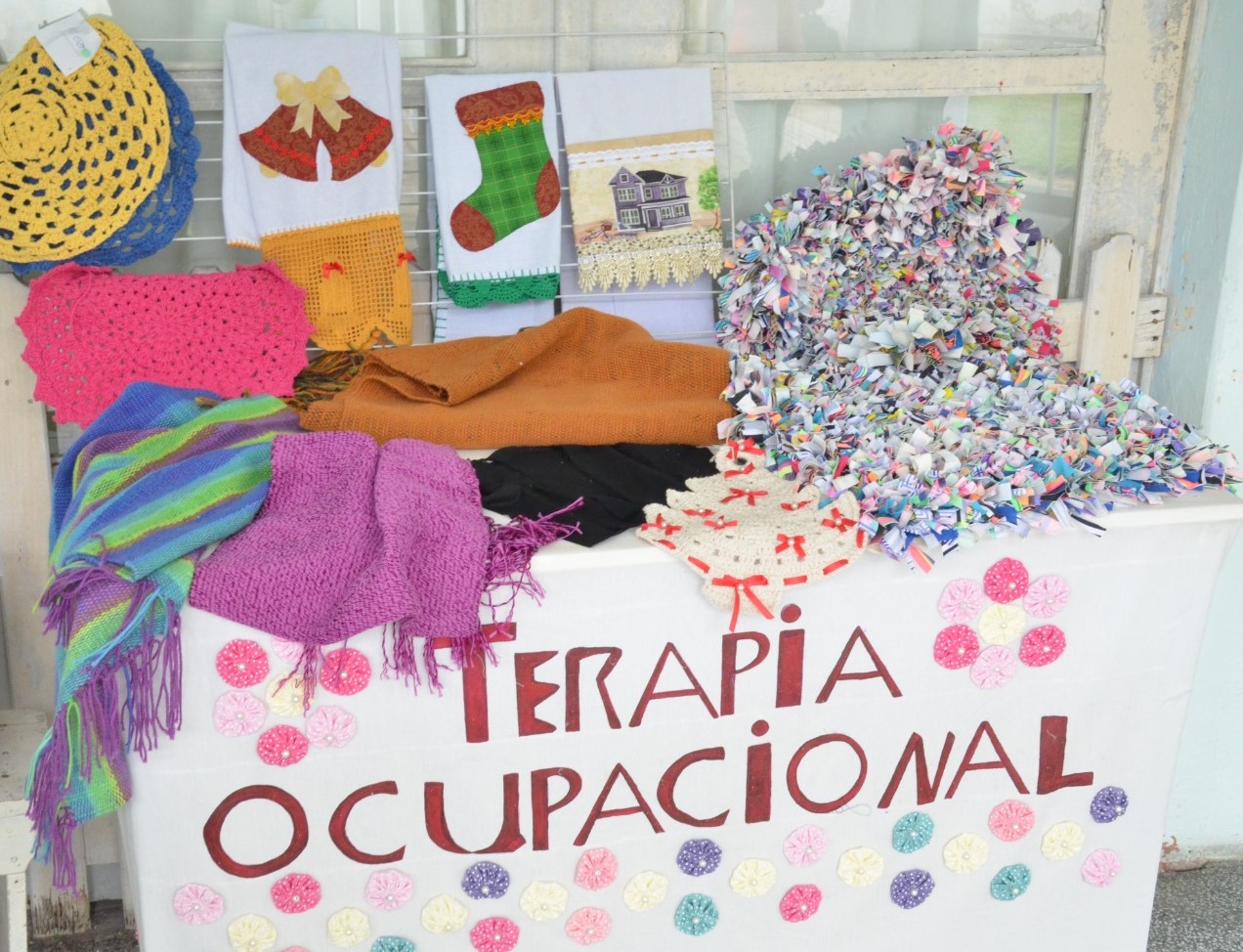 A imagem mostra uma mesa decorada com a inscrição "Terapia Ocupacional", ornada com pequenas flores coloridas de tecido. Sobre a mesa e ao fundo, há uma variedade de trabalhos manuais produzidos, como panos de prato bordados, peças de crochê, cachecóis, tapetes e artesanatos feitos com retalhos. As cores vivas e a diversidade de texturas transmitem um clima de criatividade, acolhimento e valorização do trabalho artesanal, evidenciando o papel da terapia ocupacional como ferramenta terapêutica e de expressão pessoal.