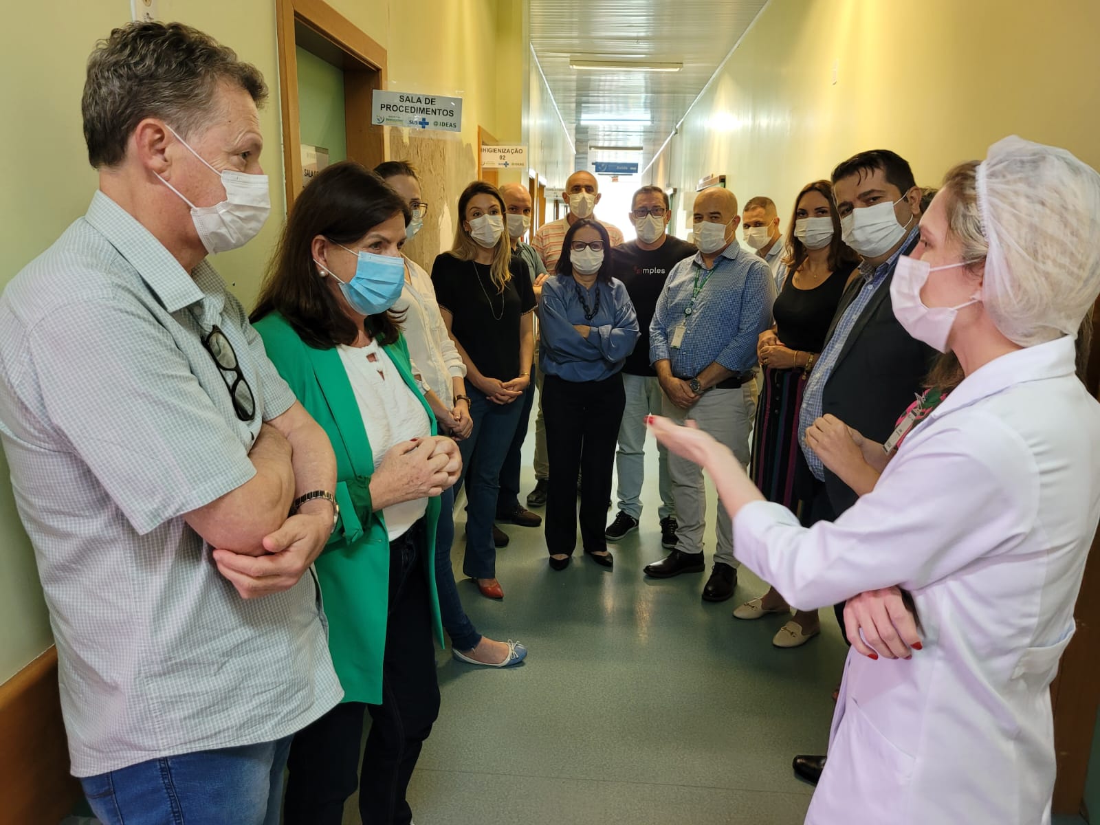 Grupo de aproximadamente 14 pessoas reunidas em um corredor de hospital, todos usando máscaras. À direita, uma profissional de saúde com jaleco branco e touca cirúrgica está falando e gesticulando, enquanto os demais escutam atentamente. Na parede há placas com nomes de salas, como “Sala de Procedimentos” e “Higienização”.