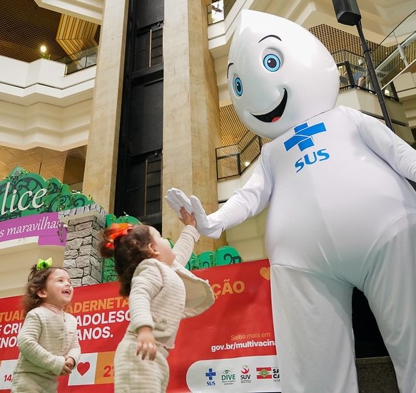 Mascote Zé Gotinha, símbolo de campanhas de vacinação do SUS, cumprimenta com um toque de mão duas meninas pequenas sorridentes, em ambiente interno decorado com faixa de divulgação da multivacinação.