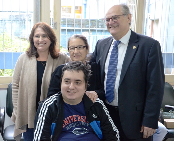 Quatro pessoas sorrindo posam para a foto em uma sala iluminada. Em destaque, um homem jovem cadeirante com camiseta azul-marinho e jaqueta preta com listras brancas. Atrás dele, duas mulheres e um homem de terno com óculos sorriem, demonstrando um momento de confraternização e alegria.