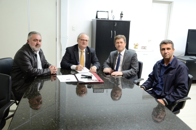 Quatro homens sentados ao redor de uma mesa de reunião. Um deles, ao centro, assina documentos com uma caneta enquanto os demais observam.