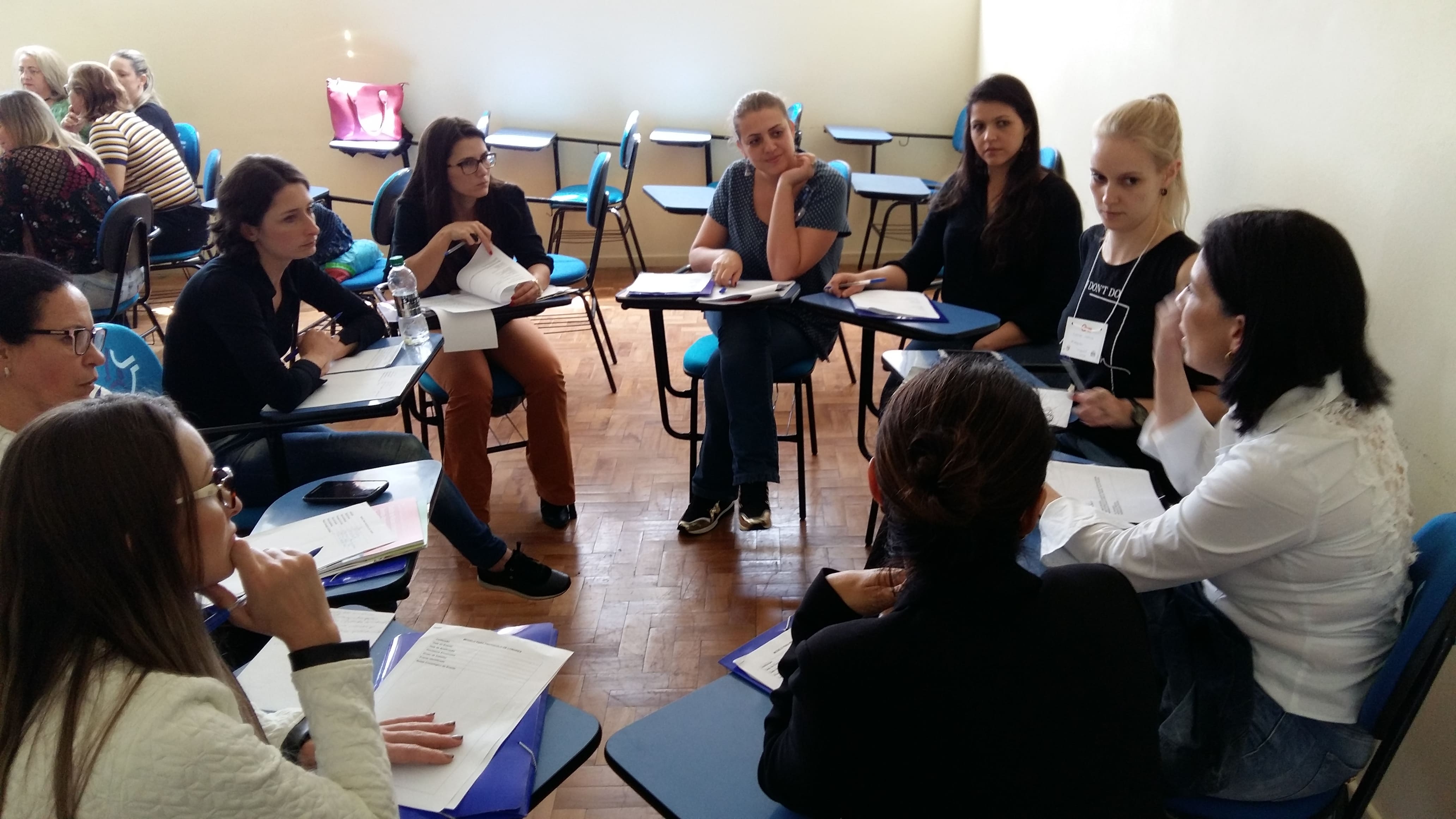 Grupo de mulheres sentadas em círculo em uma sala de aula durante uma oficina. Elas discutem documentos e anotações sobre carteiras azuis de treinamento. Algumas têm crachás, e uma das participantes fala enquanto as outras escutam atentamente. Ao fundo, outros grupos também participam da atividade em um ambiente iluminado por luz natural.