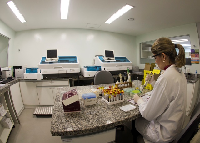 A imagem mostra uma profissional da área da saúde trabalhando em um laboratório clínico moderno. Ela veste jaleco branco, luvas e óculos de proteção, e está manuseando tubos de ensaio sobre uma bancada de granito.