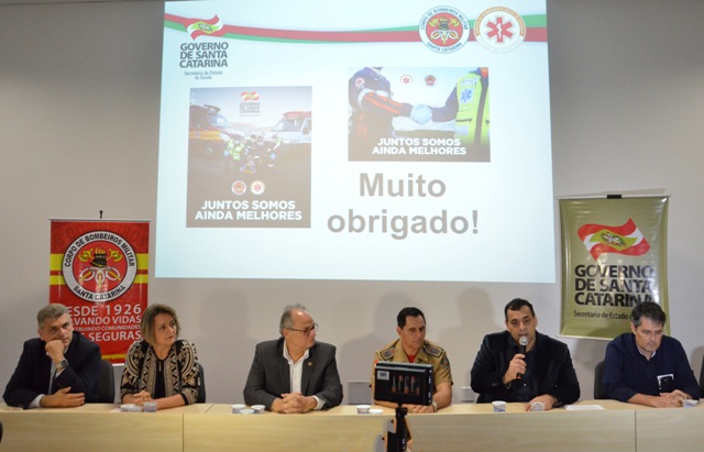 Coletiva de imprensa com autoridades do Governo de Santa Catarina e do Corpo de Bombeiros, apresentando a integração do SAMU com os Bombeiros Militares. Sete pessoas estão sentadas à mesa, com um telão ao fundo exibindo a mensagem 'Juntos somos ainda melhores' e 'Muito obrigado!'.