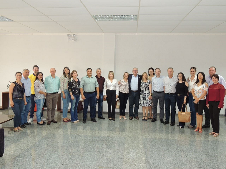 Grupo de aproximadamente 20 pessoas posando para foto em ambiente interno, de frente para uma parede clara. Todos estão em pé, lado a lado, sorrindo, vestindo roupas formais e casuais.