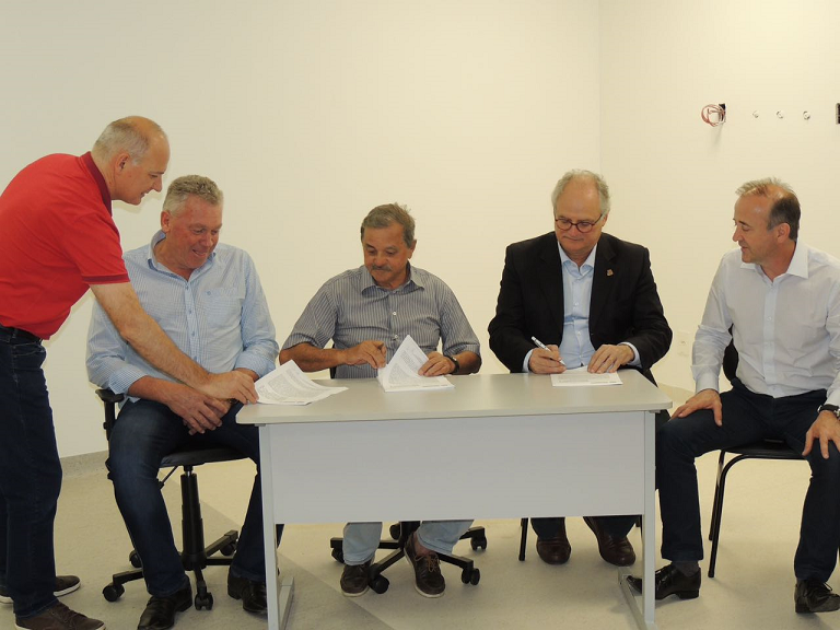 Cinco homens estão reunidos em uma sala com paredes brancas e piso claro. Três deles estão sentados atrás de uma mesa branca, enquanto dois estão de pé ao lado, interagindo com documentos.