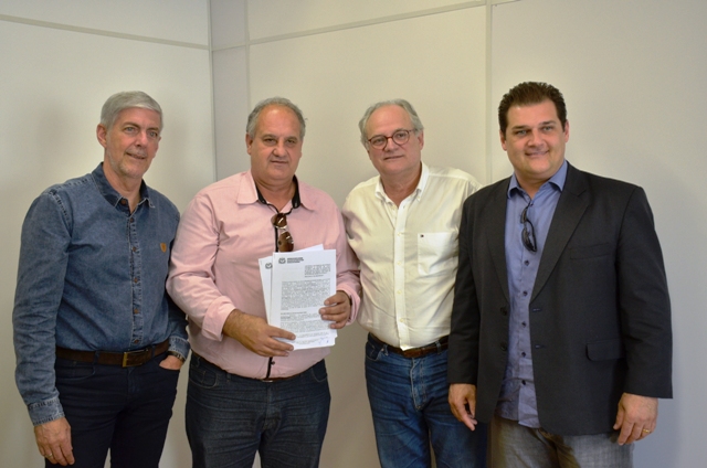 Quatro homens posam sorrindo para a foto em ambiente interno, de frente a uma parede branca. Um deles, ao centro, segura em mãos um documento assinado.