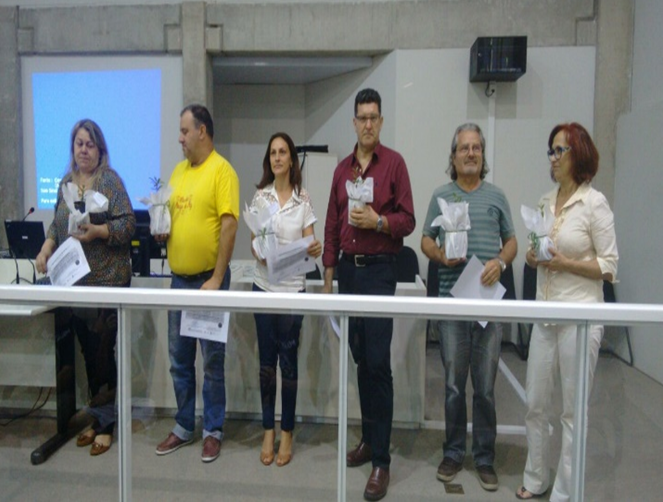 Seis pessoas recebem homenagens durante o II Encontro pela Cultura da Paz, em São José. Elas estão alinhadas em frente a uma mesa, segurando certificados e lembranças em embalagem transparente, com ambiente de auditório ao fundo.