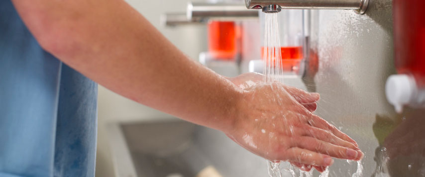 Profissional da saúde, com uniforme azul, lava cuidadosamente as mãos sob uma torneira de aço inox, com água corrente.