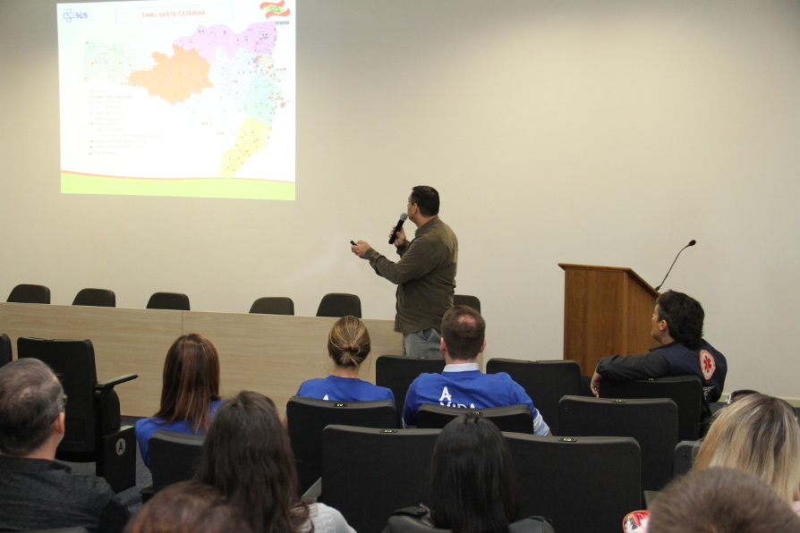 Homem em pé, de costas, aponta para mapa projetado em tela durante palestra sobre atendimento pré-hospitalar no estado de Santa Catarina. Sentados à frente, militares e profissionais de saúde acompanham atentamente, alguns usando uniformes com logos do SAMU e Corpo de Bombeiros.