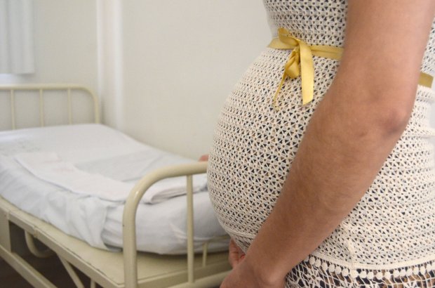 Mulher grávida de perfil, usando vestido de crochê claro com laço amarelo na barriga, em frente a um leito hospitalar. A imagem representa atenção pré-natal e cuidados com a gestação.