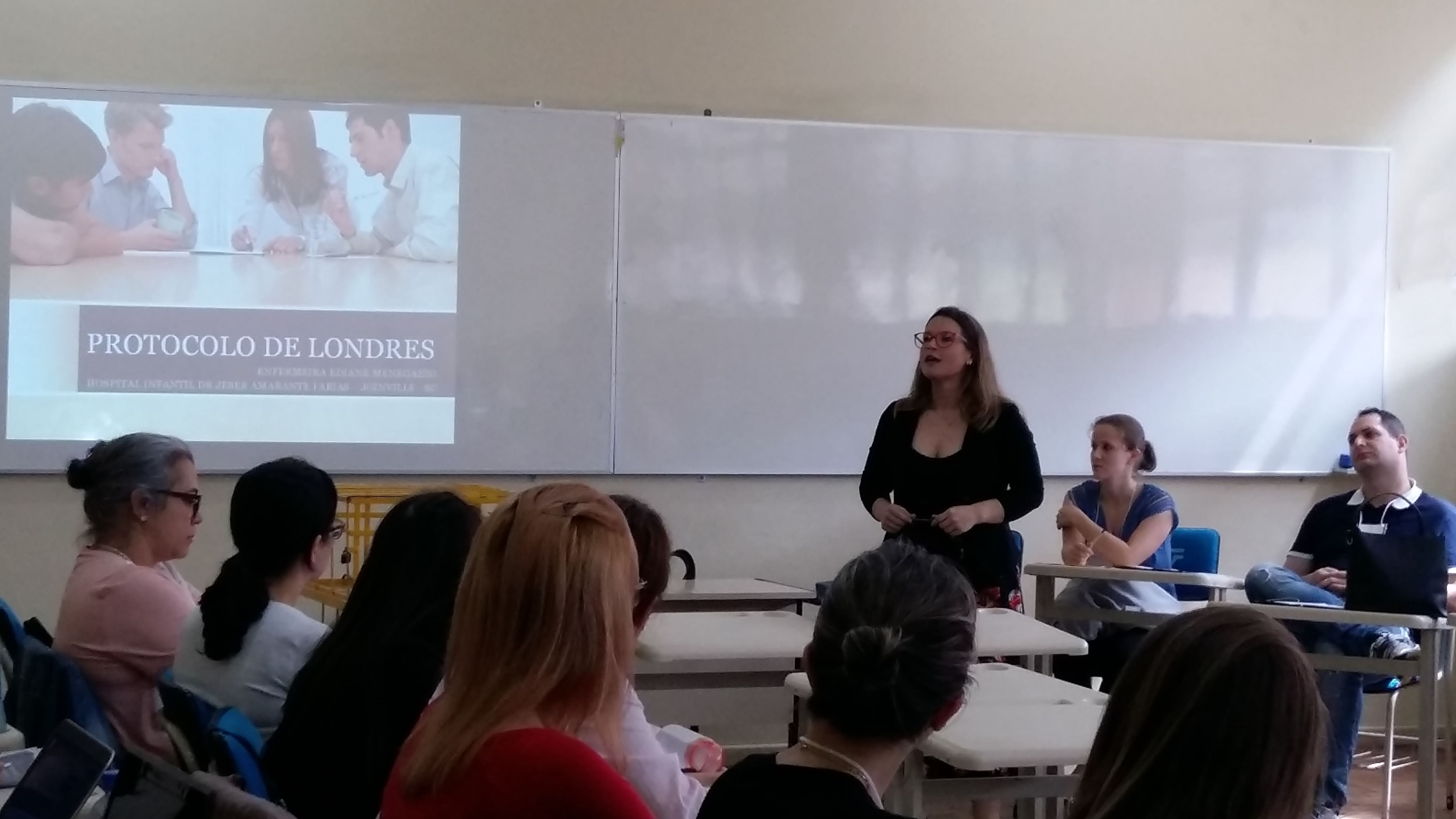 Sala de aula com uma mulher de pé, palestrando para um grupo de pessoas sentadas. 
