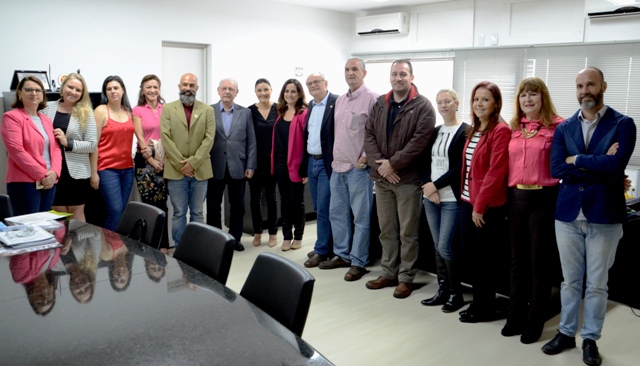 Foto de uma solenidade de posse ocorrida em uma sala de reuniões. Um grupo de cerca de 16 pessoas, entre homens e mulheres, posa sorridente para a foto, lado a lado, em frente a uma mesa de reuniões comprida.