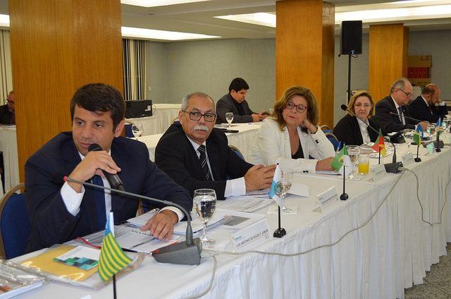 Secretários estaduais de saúde e representantes participam da 14ª Assembleia do Conselho Nacional de Secretários da Saúde (Conass), em Brasília. Os participantes estão sentados em uma mesa retangular coberta com toalha branca, com bandeiras dos estados e materiais de trabalho à frente, enquanto discutem temas da saúde pública nacional.