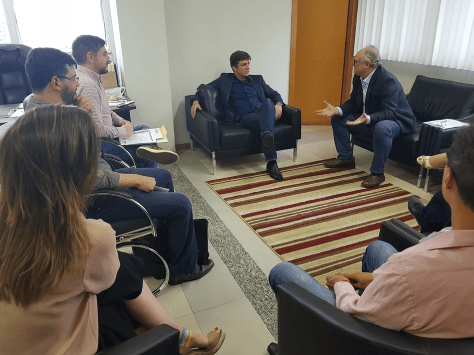 Reunião em gabinete entre o secretário de Estado da Saúde, Vicente Caropreso, e o prefeito de Balneário Camboriú, Fabrício Oliveira, acompanhados por técnicos e autoridades municipais. O encontro ocorre em ambiente formal, com todos sentados em poltronas ao redor de um tapete listrado, discutindo ações de combate ao mosquito Aedes aegypti.