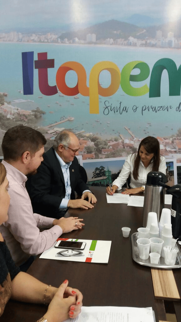 Secretário de Estado da Saúde, Vicente Caropreso, e a prefeita de Itapema, Nilza Simas, durante a assinatura de um termo de compromisso em reunião oficial. O encontro acontece em uma sala com mesa de reuniões, materiais institucionais e uma bandeja com copos e garrafa térmica.