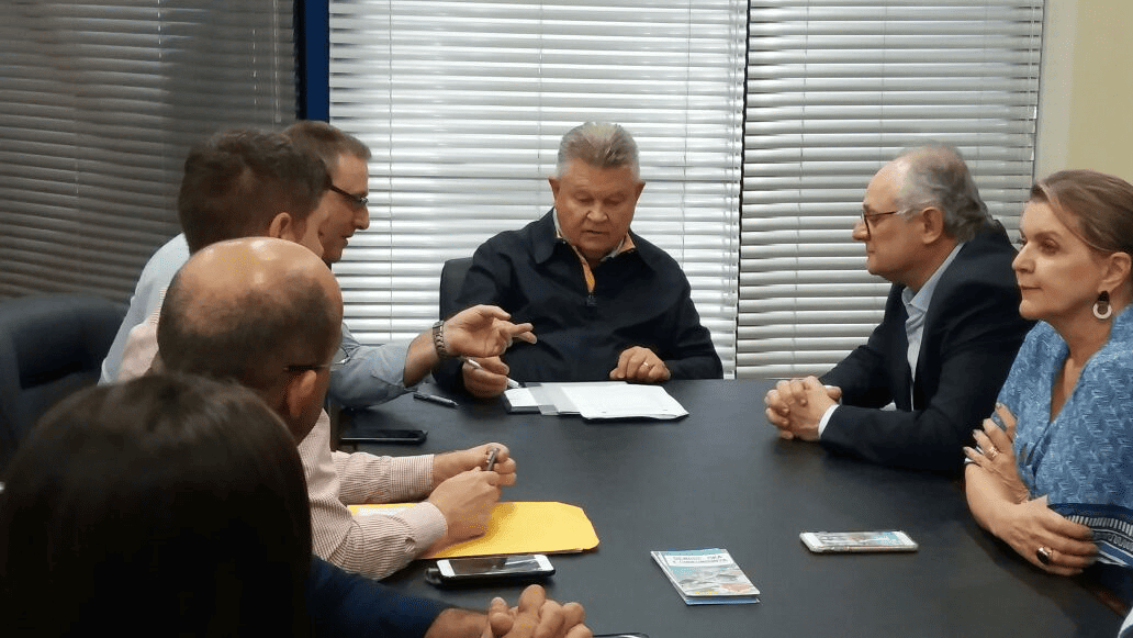 Secretário de Estado da Saúde, Vicente Caropreso, em reunião com o prefeito de Joinville, Udo Döhler, e demais autoridades municipais. Eles estão sentados ao redor de uma mesa de reuniões, discutindo documentos relacionados à saúde pública e combate ao Aedes aegypti. O ambiente é corporativo, com persianas fechadas ao fundo.