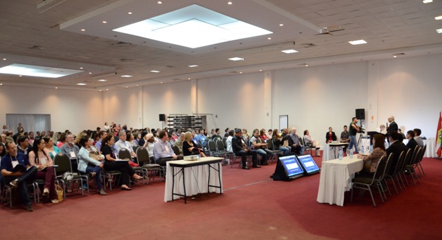 Auditório amplo com participantes sentados assistindo à abertura da 1ª Conferência de Vigilância em Saúde de Santa Catarina. À direita, uma mesa com palestrantes e telões exibindo apresentações. O evento discute propostas para a Política Nacional de Vigilância em Saúde e conta com representantes de todos os 295 municípios catarinenses.