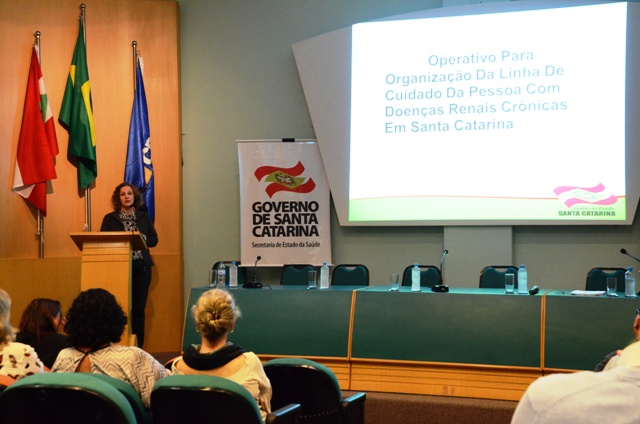 Imagem de uma apresentação oficial realizada em um auditório. À esquerda, uma mulher fala ao microfone em um púlpito. Atrás dela, estão posicionadas as bandeiras de Santa Catarina, do Brasil e outra institucional.