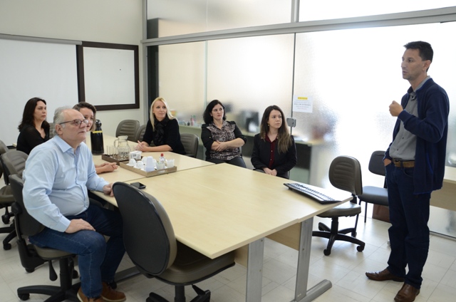 Imagem mostra uma reunião em ambiente corporativo. À esquerda, seis pessoas — quatro mulheres e dois homens — estão sentadas ao redor de uma mesa retangular de madeira clara, ouvindo atentamente um homem em pé à direita, que gesticula enquanto fala. A sala possui divisórias de vidro fosco ao fundo e mobiliário de escritório simples. Em cima da mesa, há garrafas de café, copos de vidro, teclado e mouse. A cena retrata uma reunião técnica no setor de Telemedicina do Governo de Santa Catarina.