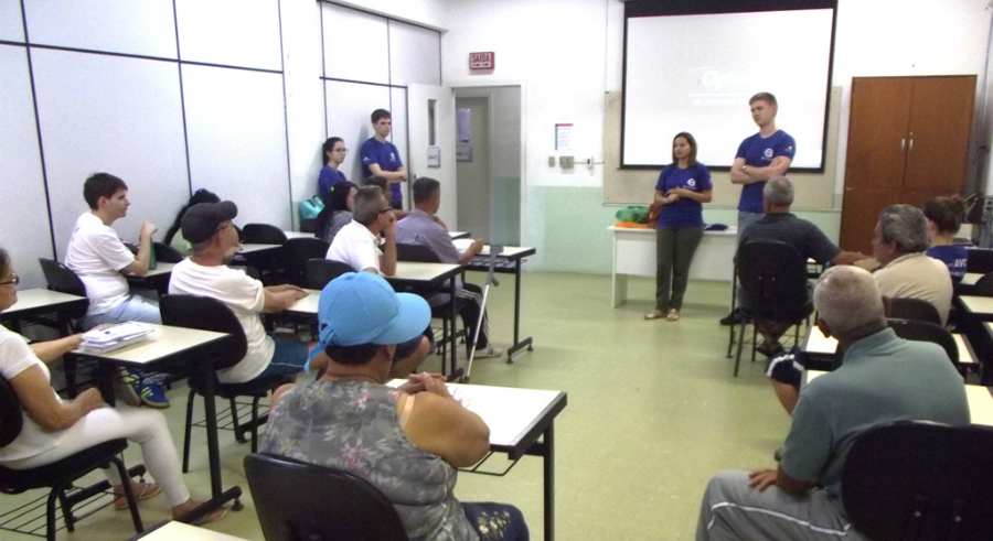 Sala de aula com participantes sentados em carteiras, ouvindo dois jovens em pé à frente, que usam camisetas azuis da Udesc e apresentam um projeto de reabilitação para pessoas com sequelas de AVC; ao fundo, uma tela de projeção apagada e uma terceira integrante da equipe em pé próxima à porta.
