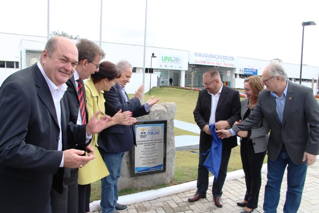 Autoridades políticas e gestores da saúde participam de uma cerimônia de inauguração em frente ao novo Centro Integrado de Saúde (CIS) de Itajaí, Santa Catarina. Eles estão reunidos ao redor de uma placa inaugural fixada em um pedestal de pedra, alguns aplaudindo e outros realizando o ato simbólico de descerramento com uma bandeira azul.