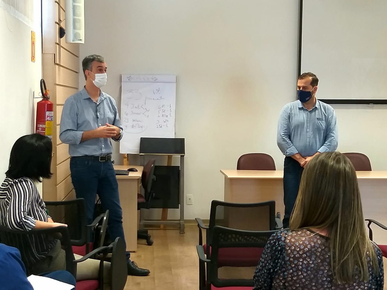 Reunião em ambiente interno com paredes claras e móveis de escritório. Dois homens em pé, usando máscaras, camisas sociais e calças jeans, falam para um pequeno grupo sentado. Ao fundo, há um flip chart com anotações feitas à mão, uma mesa com cadeiras e um projetor. Um extintor de incêndio está fixado na parede à esquerda. Participantes da reunião observam atentamente, entre eles mulheres sentadas nas primeiras fileiras.