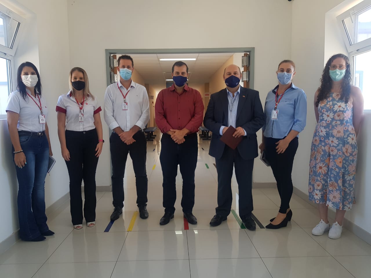 Oito pessoas, quatro homens e quatro mulheres, posam lado a lado em um corredor iluminado, todas usando máscaras. Os homens usam camisas sociais e calças formais, enquanto as mulheres vestem roupas sociais e uma delas está com vestido floral. Ao fundo, uma porta aberta revela parte de uma sala.