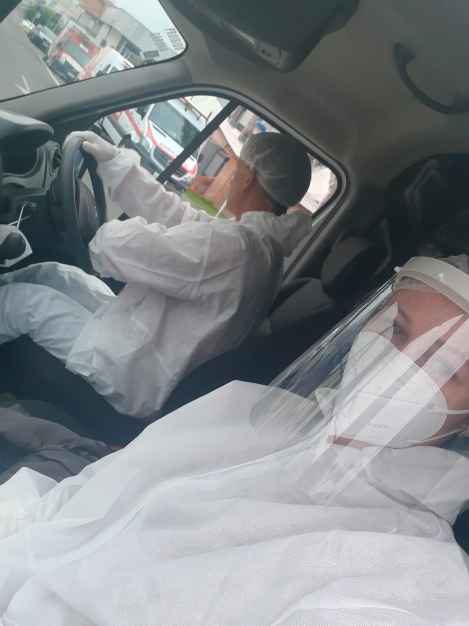 Dois profissionais da saúde, com máscaras e protetores faciais transparentes, posam no interior de um veículo.