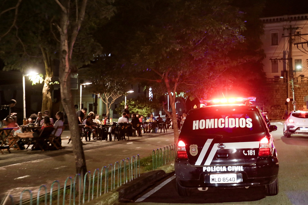 Viatura policial em operação de fiscalização circulando por rua comercial com bares abertos e pessoas sentadas nas mesas externas, durante monitoramento das medidas restritivas em Santa Catarina.