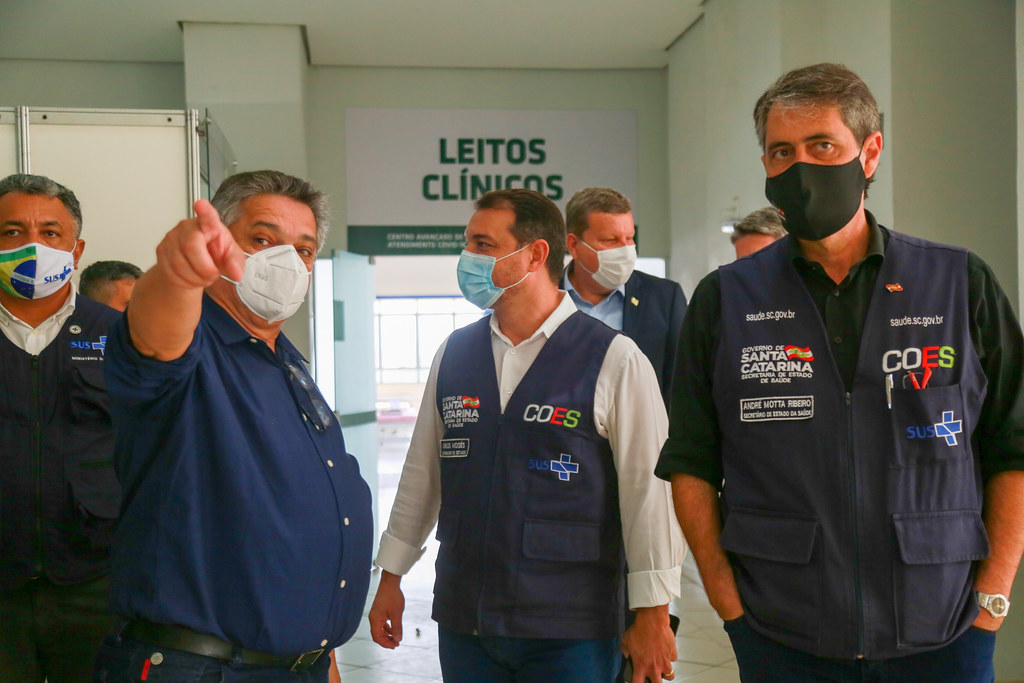 Três homens usando máscara; o da direita aponta para a frente. Ao fundo, uma sala com placa identificando 'Leitos Clínicos'.
