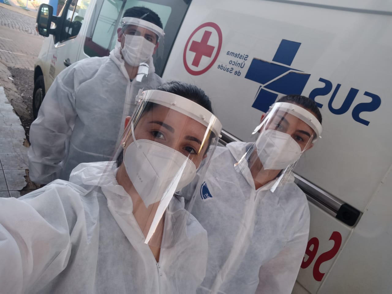 Três profissionais da saúde tiram selfie usando máscaras e protetores faciais transparentes, posicionados em frente a uma ambulância do SUS.