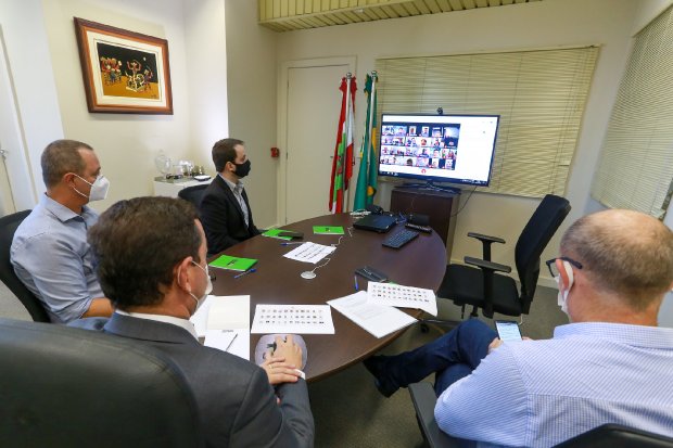 Quatro homens sentados à mesa em uma reunião, com uma tela ao fundo exibindo uma videochamada.