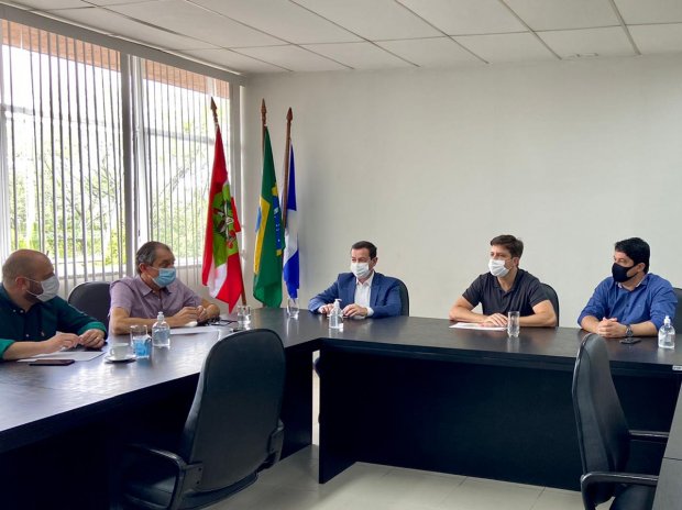 Seis homens usando máscaras estão reunidos em uma sala de reuniões, sentados ao redor de duas mesas pretas em formato de L. Ao fundo, há três bandeiras: a de Santa Catarina, do Brasil e uma azul e branca. A sala tem paredes brancas e uma janela grande com persianas, permitindo a entrada de luz natural.