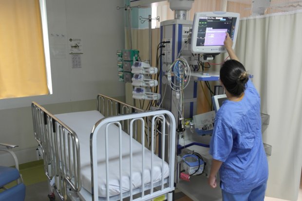 Profissional de saúde com uniforme azul opera um monitor hospitalar ao lado de um leito vazio com grades metálicas em um quarto de internação. Equipamentos médicos estão conectados ao monitor, e o ambiente é bem iluminado, com cortinas claras ao fundo.