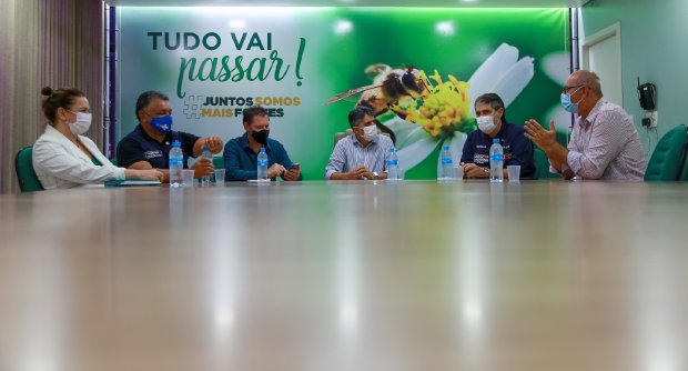 Seis pessoas sentadas à mesa, cada uma com uma garrafa de água à frente; ao fundo, cartaz com os dizeres 'Tudo vai passar! Juntos somos mais fortes'.