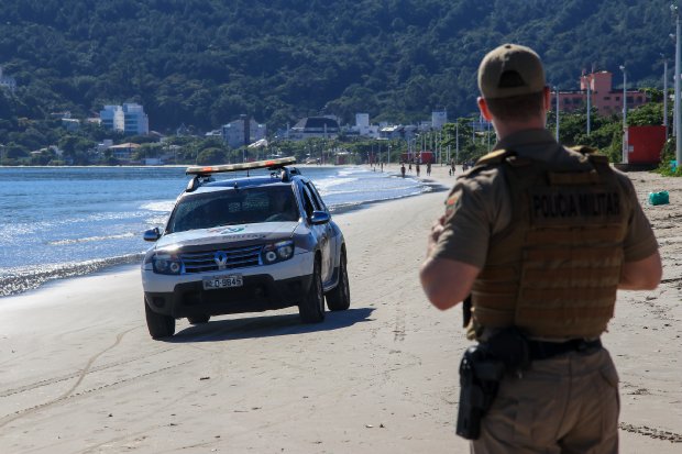 Policial masculino de costas observa sua viatura estacionada na beira da praia, com o mar ao fundo.