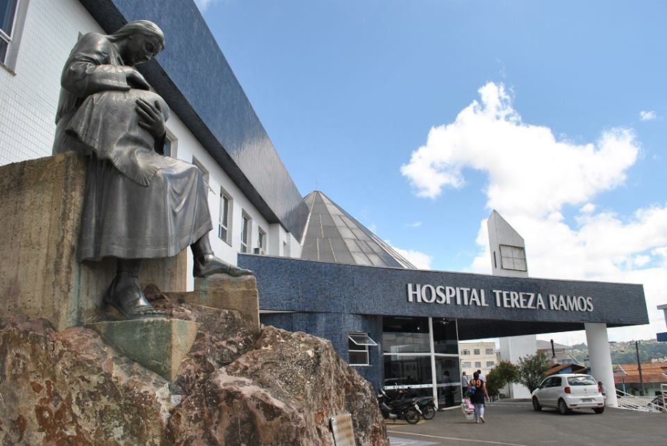Estátua em primeiro plano com a fachada do Hospital Tereza Ramos ao fundo, durante anúncio da retomada das obras e ampliação de leitos de UTI em Lages.