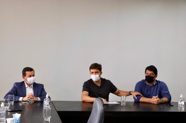 Três homens usando máscaras estão sentados lado a lado em uma mesa de reunião preta. O homem do centro, com camiseta preta, toca no ombro do homem à direita, que veste camisa azul. O terceiro homem, à esquerda, veste terno azul e camisa branca. Sobre a mesa, há copos, garrafas de água e um frasco de álcool em gel. O fundo é uma parede branca lisa.