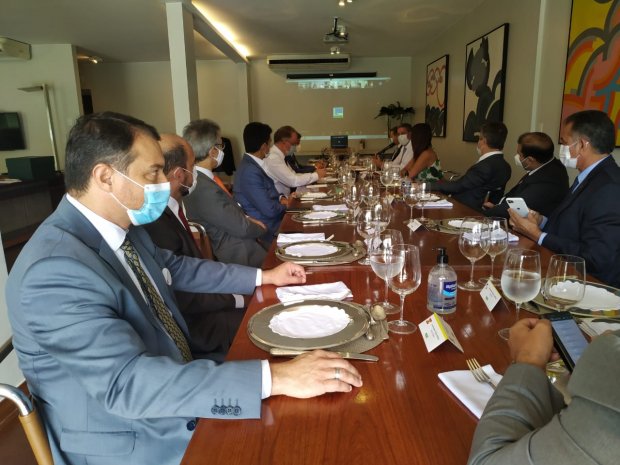 Grupo de pessoas, todas usando máscara, sentadas ao redor de uma mesa de jantar longa e bem posta, com pratos, talheres, taças e guardanapos organizados. A reunião acontece em ambiente fechado, moderno e iluminado. Ao fundo, há um projetor exibindo conteúdo em uma tela. Alguns participantes observam a projeção, enquanto outros conversam ou utilizam o celular.