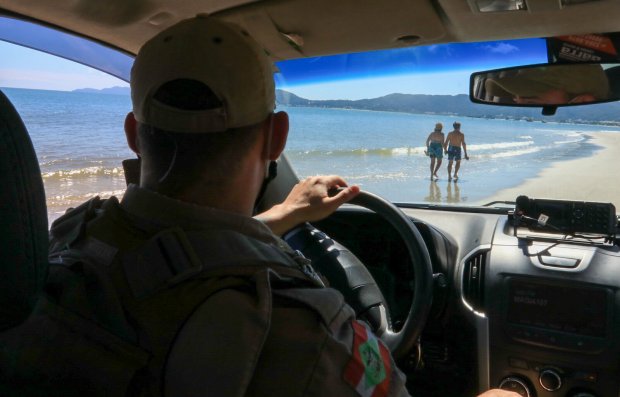 Policial dentro de uma viatura patrulha a orla da praia; à frente, duas pessoas caminham de mãos dadas, mantendo boa distância.