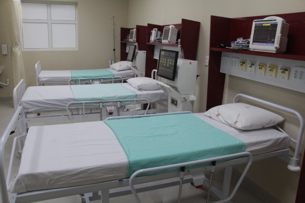 A imagem mostra uma unidade hospitalar com leitos organizados e prontos para uso. Há três camas hospitalares, todas equipadas com lençóis brancos e cobertores verdes claros, monitores multiparâmetros instalados sobre cada leito, e estrutura de suporte de equipamentos médicos fixados na parede.  Este ambiente parece ser uma UTI (Unidade de Terapia Intensiva) ou sala de internação preparada para receber pacientes, possivelmente em contexto de pandemia, como a COVID-19, dada a configuração de isolamento e monitoramento individualizado. O espaço está limpo, iluminado e pronto para atendimento clínico.