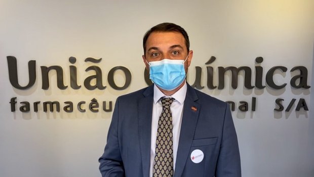 Homem usando terno azul e máscara cirúrgica posa em frente à parede com o logotipo da empresa 'União Química Farmacêutica Nacional S/A'. Ele está usando um crachá redondo no paletó e olha diretamente para a câmera.