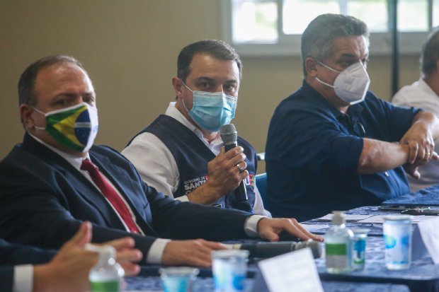Foto do governador de Santa Catarina segurando um microfone, acompanhado de dois homens sentados à mesa.