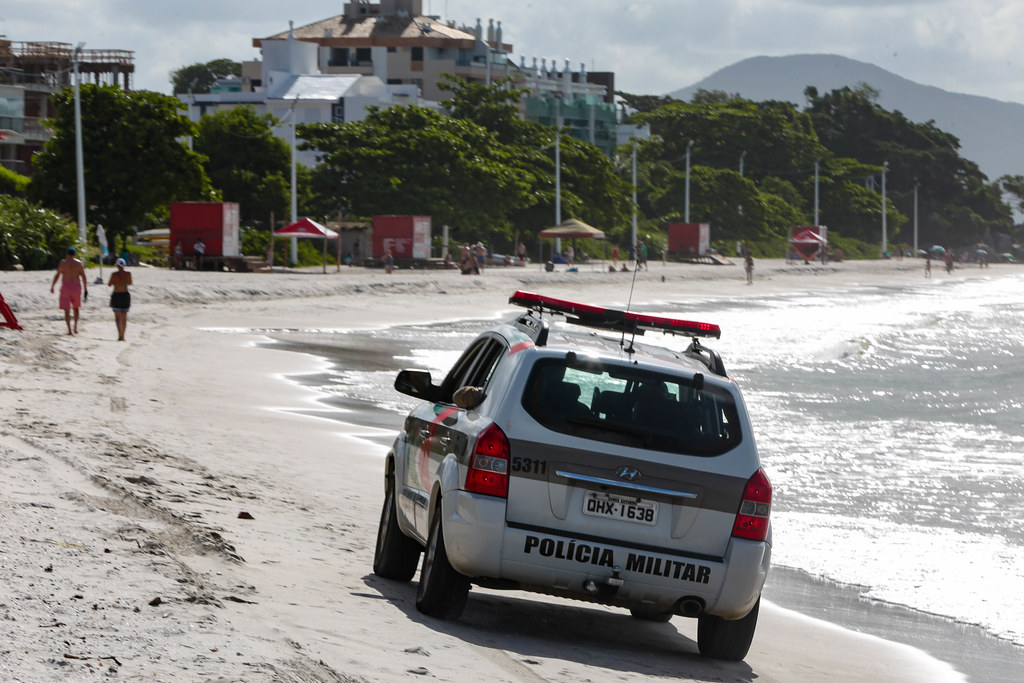 Viatura da Polícia Militar circula pela faixa de areia de uma praia durante o dia, com o mar à direita e prédios residenciais ao fundo. Algumas pessoas caminham na praia, enquanto outras estão sob guarda-sóis. A ação policial aparenta fazer parte de patrulhamento rotineiro ou fiscalização em área litorânea.