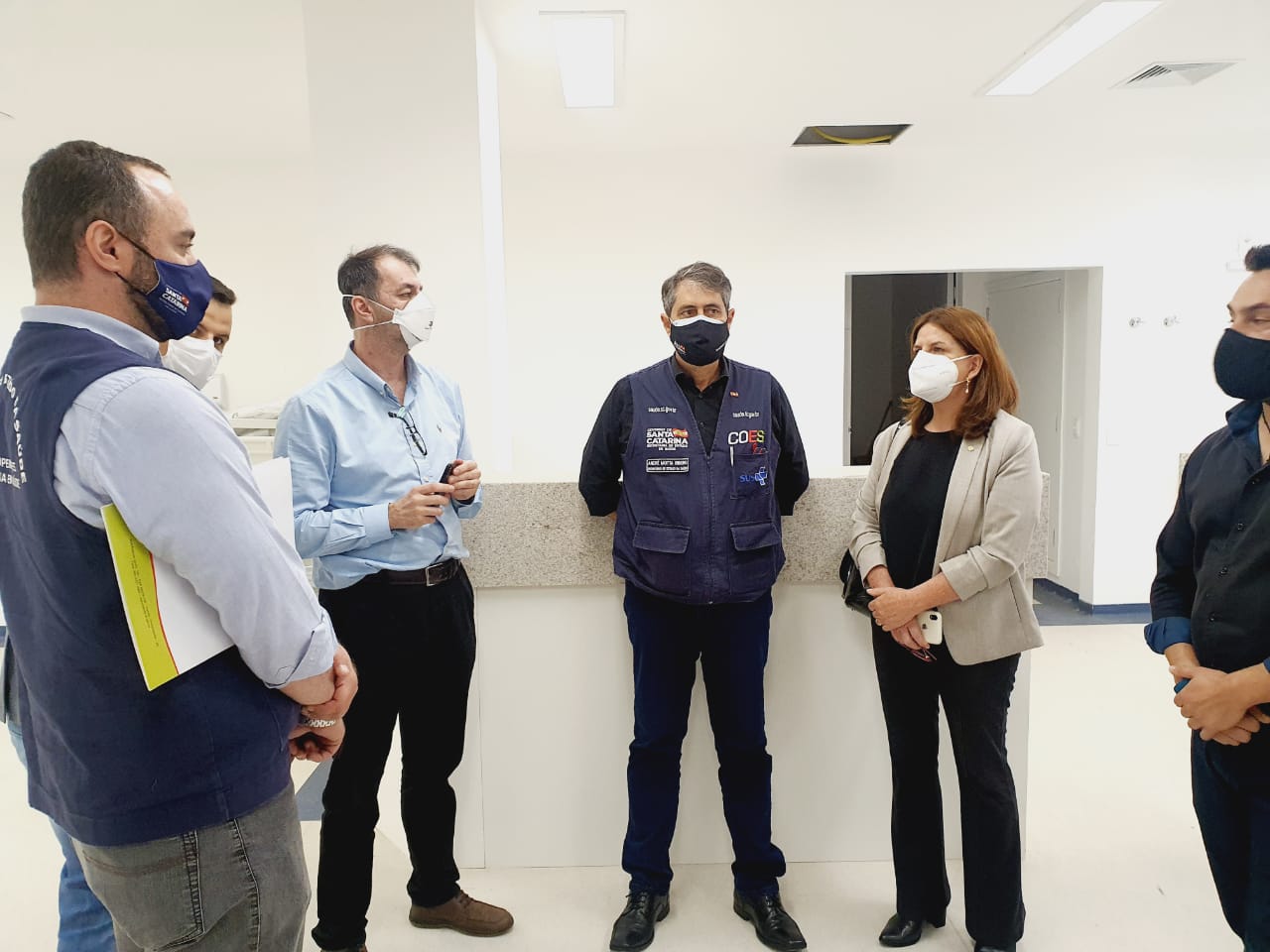 Secretário de Saúde de Santa Catarina em reunião com cinco profissionais na Serra Catarinense, todos em pé em sala de reuniões, durante agenda oficial.