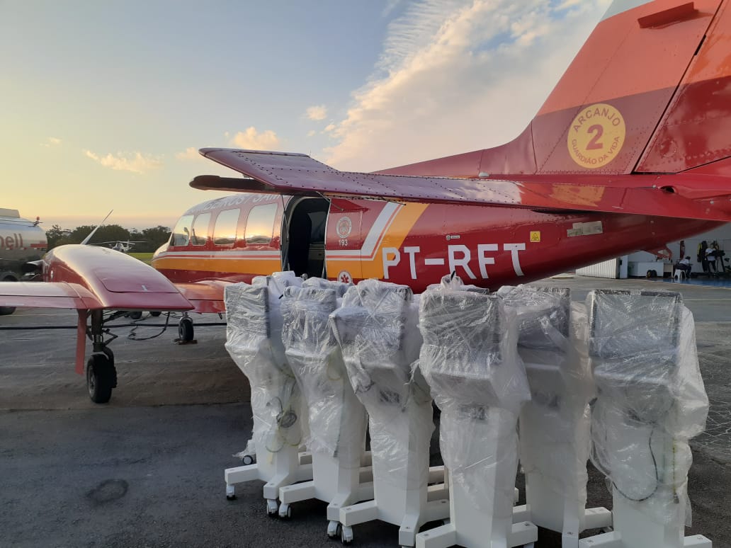 Avião vermelho e amarelo do Arcanjo 2, com porta aberta, estacionado em pista de aeroporto ao entardecer. Em frente à aeronave, vários ventiladores pulmonares embalados em plástico aguardam transporte.