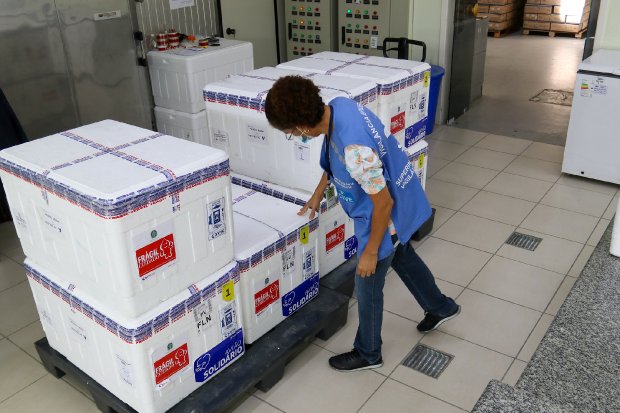 Funcionária da saúde, com colete azul e máscara, inspeciona caixas térmicas brancas empilhadas em um pallet, contendo vacinas armazenadas em centro de distribuição.