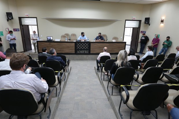 Auditório com cadeiras organizadas respeitando o distanciamento social, onde ocorre uma reunião com pessoas sentadas e outras em pé. Na mesa central, dois homens falam ao microfone em frente ao letreiro 'AMFRI'.